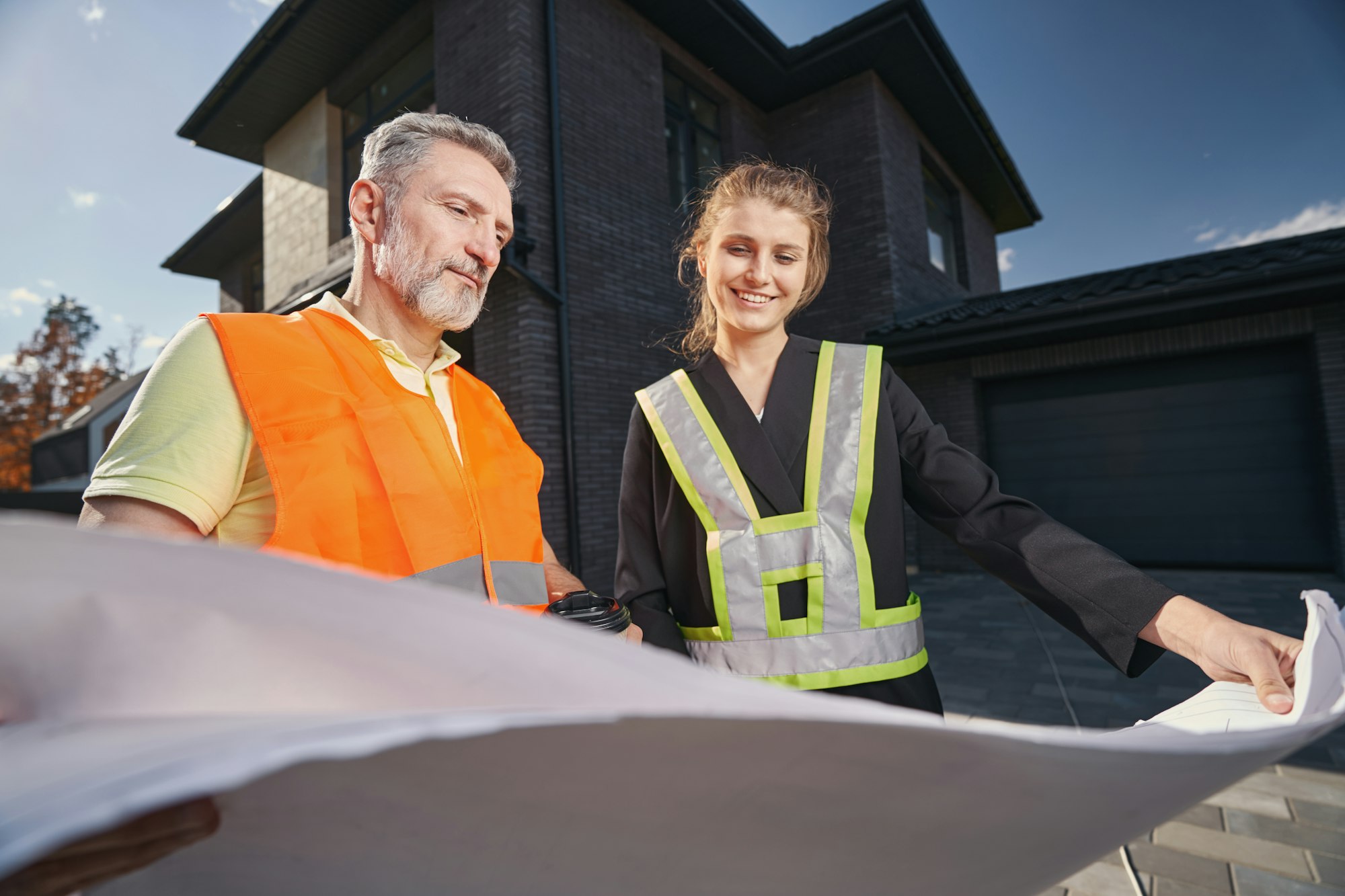 Aging builder and woman in construction vests staring at blueprints